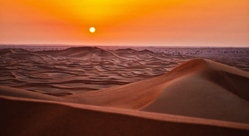 Sandscape of a desert with rolling dunes as the sun sets, turning the sky yellow and orange, and the sand purple